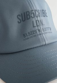 Căciulă gri dintr-un material neted. Prezintă o visieră curbată, detalii cusute și text imprimat pe față și pe lateral.