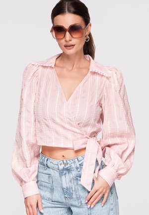 Femme portant de grandes lunettes de soleil marron, un chemisier rose texturé cache-cœur à manches bouffantes et un jean bleu clair taille haute.