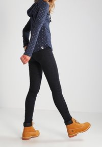 Persoon die loopt in een marineblauwe gebreide hoodie, zwarte skinny jeans en tan kleurige veterschoenen tegen een effen lichte achtergrond.