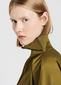 Indumento verde oliva con colletto alto e design a pieghe. Texture liscia e una leggera lucentezza. Presenta un dettaglio con zip sul collo.