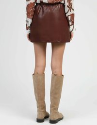 Une personne portant une mini-jupe en cuir marron, un chemisier à manches longues à motif paisley et des bottes en daim beiges haute jusqu'au genou, se tenant devant un fond uni.