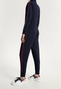 Tuta sportiva blu navy con strisce rosse sui lati, caratterizzata da un colletto alto e polsini a costine, abbinata a sneakers bianche e rosse. Tessuto con texture liscia.