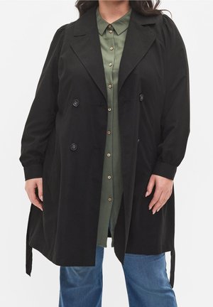 Person iført en lang sort dobbeltradet frakke over en olivengrøn skjorte med knapper og blå jeans, stående mod en ensfarvet baggrund.