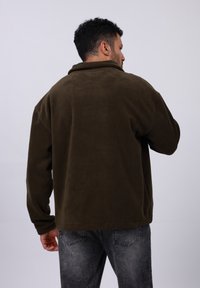 Veste en polaire marron avec un col, coupe décontractée et manches longues. Le modèle porte un jean gris foncé. L'arrière-plan est entièrement blanc.