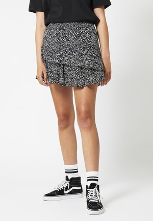 Vrouw draagt een zwart-witte geplooide minirok met patroon, een zwart T-shirt, witte sokken met zwarte strepen en zwarte hoge sneakers.