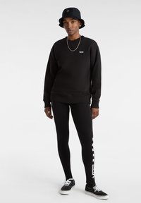 Felpa nera con logo "VANS", abbinata a leggings neri con un design bianco sul lato e sneakers nere. Un cappello a secchiello completa il look.