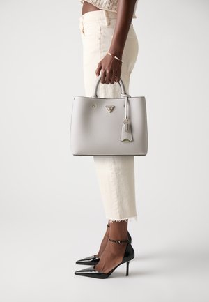Graue Lederhandtasche mit strukturierter Form, goldenen Hardware-Akzenten und einem abnehmbaren Anhänger, gehalten von einer Person in beigen Hosen und schwarzen High Heels.