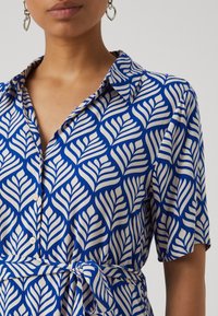 Blue shirt featuring a leaf pattern in white, with a collared design, short sleeves, and a tied waist detail. Smooth texture.