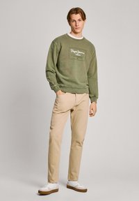 Sudadera verde oliva con cuello redondo y parche de logo en el frente, combinada con pantalones beige y zapatillas blancas con suelas marrones.