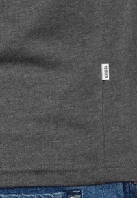 Graues Baumwoll-T-Shirt mit glattem Material, Seitennaht mit einem kleinen weißen Etikett mit der Aufschrift „!SOLID“ und kontrastierendem blauen Denim am Saum.