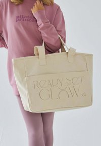 Beżowa torba na ramię z płótna z napisem "READY SET GLOW" w jasnym, wytłoczonym kroju pisma, z dwoma płaskimi uchwytami i sztywnym prostokątnym kształtem.