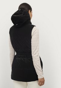 Gilet imbottito nero con colletto alto e cappuccio con cordino. Presenta una vita con cintura e una consistenza liscia, abbinato a un top a maniche lunghe a fantasia.