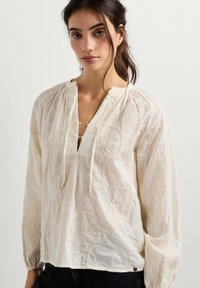 Femme aux cheveux foncés portant un chemisier ample de couleur crème avec broderies florales et un col fendu avec liens.