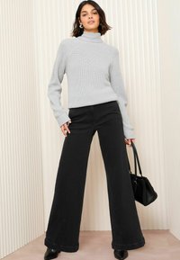 Grauer gerippter Rollkragenpullover kombiniert mit weit geschnittenen schwarzen Jeans. Das Model hält eine schwarze Handtasche und steht vor einem strukturierten hellen Hintergrund.