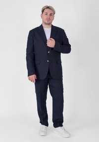 Completo in lino blu navy con giacca a due bottoni, abbinato a pantaloni coordinati e sneakers bianche. Tessuto liscio, vestibilità su misura, senza fantasia.