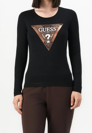Frau trägt ein schwarzes Langarmshirt mit einem großen braunen dreieckigen Guess-Logo und dunkelbraunen Hosen vor weißem Hintergrund.
