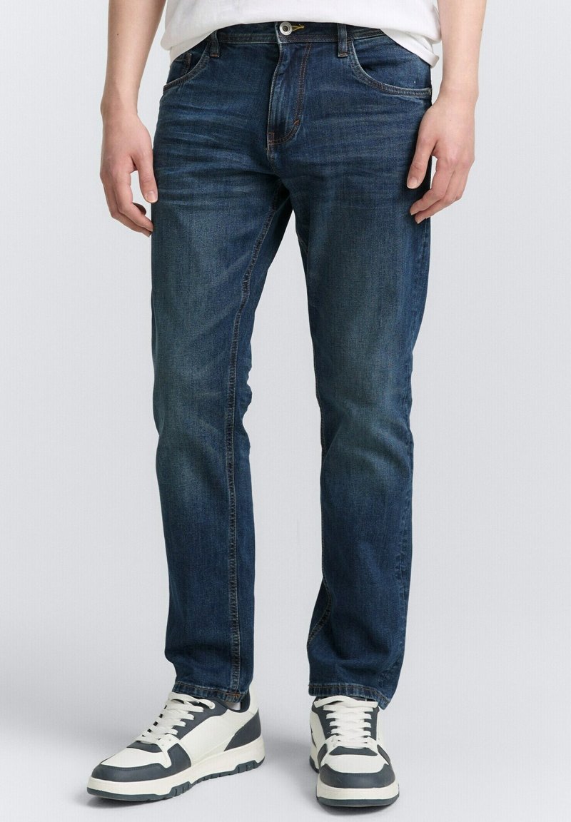 Homme portant un jean bleu foncé et des baskets blanches avec des détails noirs, debout devant un fond clair uni.
