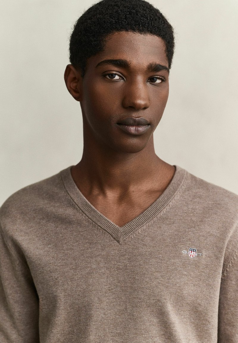 Joven con pelo rizado y corto que lleva un jersey beige de cuello en V con un pequeño logo en el pecho izquierdo, mirando hacia adelante.