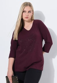 Burgunderfarbendes V-Ausschnitt-Shirt mit dreiviertel Ärmeln, das ein strukturiertes Design mit Strasssteinen aufweist. Kombiniert mit schwarzen Hosen und einer dekorativen Clutch.