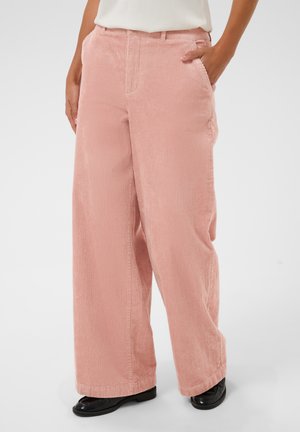 Personne portant un pantalon en velours côtelé rose ample et des chaussures noires, une main dans la poche, debout devant un fond uni.