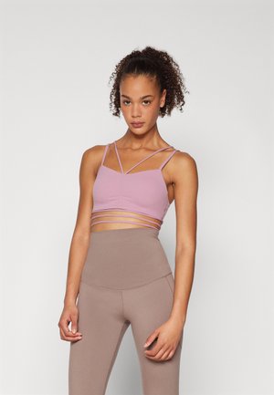 Lys rosa sportbh med flere tynne stroppar, med en glatt tekstur og en tettsittende design. Kåret med brune høyt midje leggings.