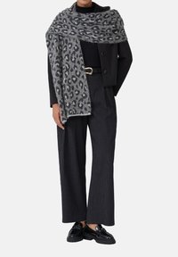 Écharpe à imprimé léopard gris drapée sur une veste noire, associée à un pantalon large noir texturé et des chaussures noires brillantes.