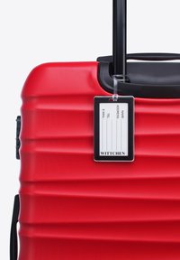 Maleta rígida roja con un acabado texturizado, que cuenta con un mango retráctil y una etiqueta de equipaje que muestra información de contacto.