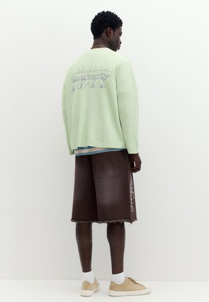 Homme portant un pull vert clair avec le texte "community" et un short marron avec des baskets, debout de dos sur fond blanc.