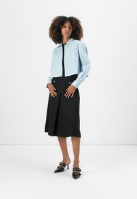 Lichtblauwe cropped jacket met een kraag, knoopsluiting en twee borstzakken, gedragen met een zwarte rok tot de knie en zwarte hakken.