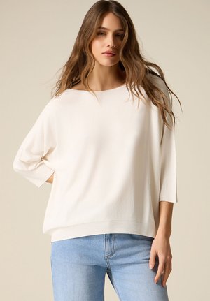 Ragazza giovane con lunghi capelli castani ondulati, indossa un maglione bianco largo con maniche a tre quarti e jeans azzurri, in piedi davanti a uno sfondo semplice.