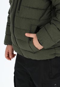 Giacca a vento verde oliva con texture trapuntata, chiusura con zip e bottoni a pressione, tasche laterali, indossata sopra pantaloni neri, mani nelle tasche.