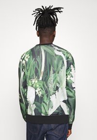 Svart bomberjacka med livfull grön och vit blommönster, med ribbad krage och muddar. Slät tygtextur, enkel skärning.