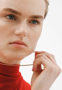 Collar de metal dorado sostenido por una mano, llevado por una persona con un jersey de cuello alto rojo. Primer plano de las características faciales, incluyendo ojos azules y piel suave.