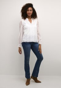 White lace blouse with geometric patterns, balloon sleeves, and a V-neckline, paired with dark blue jeans and brown heeled ankle boots.