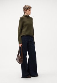 Maglione a coste di colore verde oliva con collo alto, abbinato a jeans in denim blu scuro a gamba larga. Borsa a tracolla in pelle marrone tenuta a mano.
