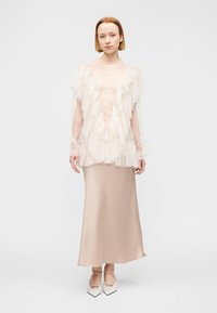 Γυναίκα που φοράει ένα διαφανές τοπ με βολάν πάνω από ένα μπεζ σατέν φόρεμα, συνδυασμένο με λευκά μυτερά τακούνια με λουράκια στον αστράγαλο.