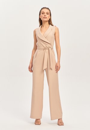Kobieta stojąca, ubrana w beżowy bezrękawnikowy kombinezon z szerokimi nogawkami i przewiązanym paskiem w talii, pozująca na jednolitym jasnym tle.