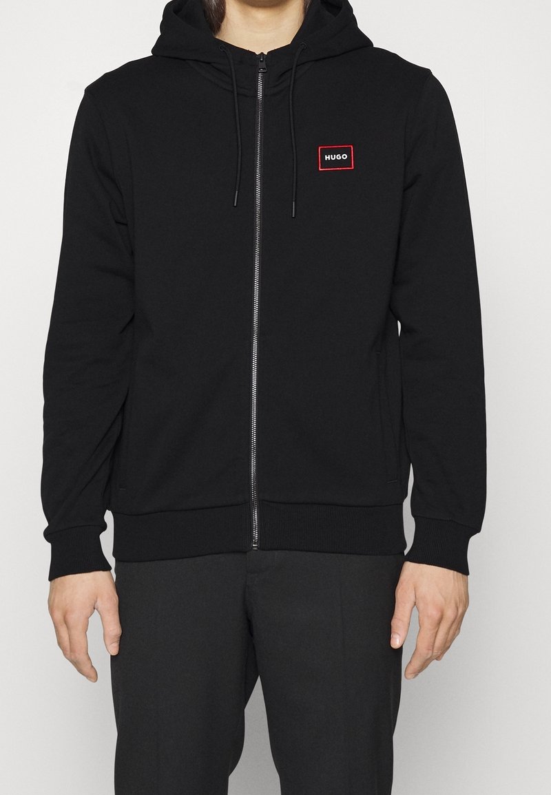 Homme portant un sweat à capuche noir zippé avec cordons et un petit logo "HUGO" rouge et blanc sur le côté gauche de la poitrine, assorti à un pantalon noir.