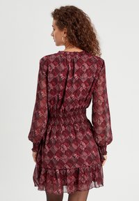 Robe à manches courtes, longueur genou dans un tissu bordeaux et noir à motifs, dotée d'une taille smockée et d'un ourlet à volants. Texture légère et transparente.
