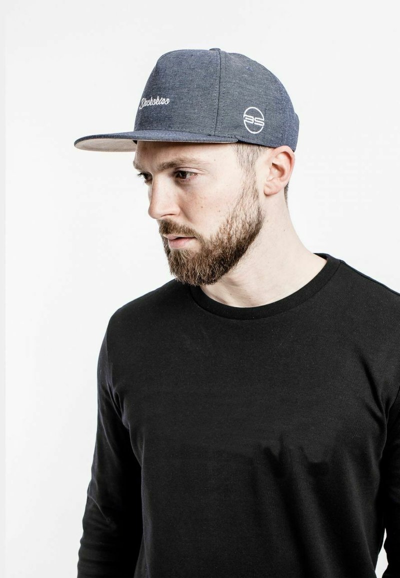 Gorra de béisbol con textura similar al denim, color azul, visera plana y logo de marca bordado en blanco. Camisa negra de manga larga llevada debajo.