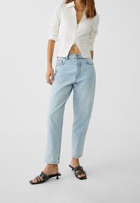 Camisa blanca ajustada con cremallera y mangas largas, jeans de talle alto de color azul claro, y sandalias de tacón abiertas de color negro con tiras decorativas.