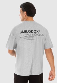 Grå bomulds-t-shirt med korte ærmer, der har fed sort tekst på ryggen: "SMILODOX TRAINING CLUB" og yderligere motiverende sætninger.
