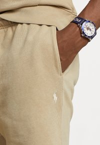 Beige bomullsbyxor med sidfickor och en liten vit broderad logga. I sällskap med en blå klocka med vit urtavla.