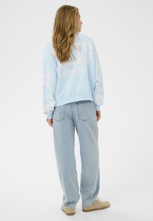 Femme aux cheveux longs portant un sweat-shirt bleu clair avec un grand texte blanc et un jean bleu clair ample, debout devant un fond blanc uni.