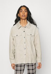 Chaqueta de pana color beige claro con cuello, dos bolsillos en el pecho, botones plateados y un corte relajado. La lleva puesta sobre pantalones de patrón de cuadros.