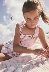 Costum de baie roz cu imprimeu floral, cu bretele volanate, așezat pe o pătură texturată roz deschis, înconjurat de diverse scoici.