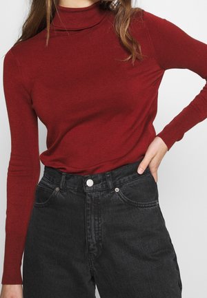 Frau trägt einen eng anliegenden roten Rollkragenpullover, der in hoch taillierte schwarze Denim-Jeans gesteckt ist, mit der Hand auf der Hüfte.