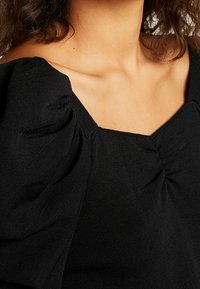 Zwarte gebreide top met een vierkante halslijn, geplooide puffmouwen en een gestructureerde afwerking, die de zachtheid en de val van de stof benadrukt.