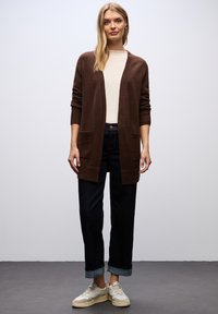 Brown knitted cardigan with open front, ribbed cuffs, and pockets; paired with cream top and dark denim jeans; worn with beige sneakers.