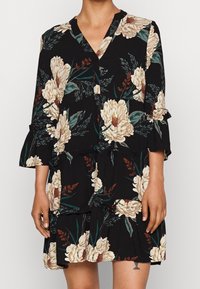 Svart blommig klänning med stora beige blommor och gröna blad, långa ärmar med volanger och en avslappnad passform med en knytning vid midjan.